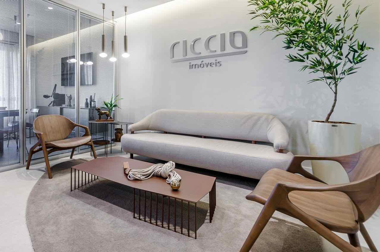Recepção da Riccio Imóveis, com o logo na parede, sofá e poltronas de madeira